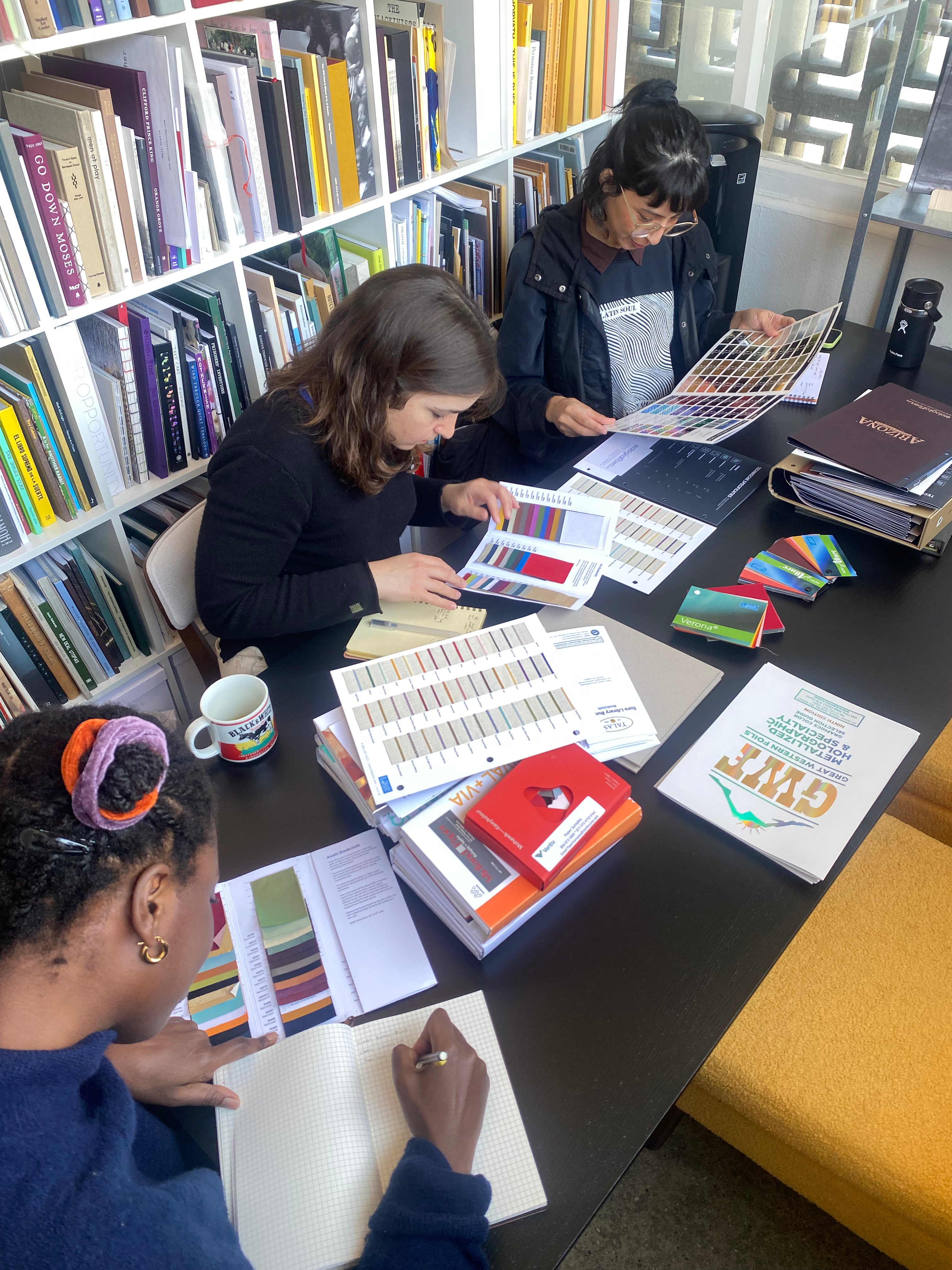 Three people working together at a table with books and color swatches in a library setting.